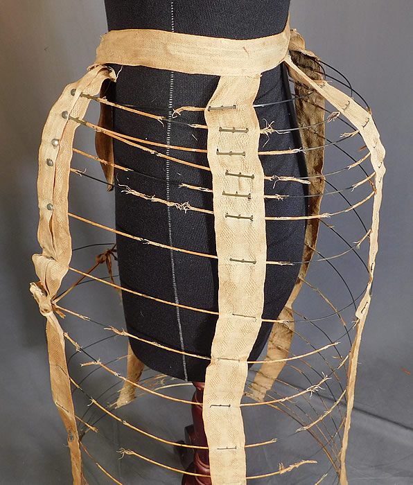 Victorian Bustle Back Wire Hoop Crinoline Cage Petticoat Skirt Support
This wonderful womans collapsible cage petticoat has a protruding bustle hump on back and an adjustable waistband strap with buckle clasp closure on the front. This unique undergarment is a type of framework that would have been worn under a bustle dress to expand the fullness and support the back drape of the fanned train skirt.
