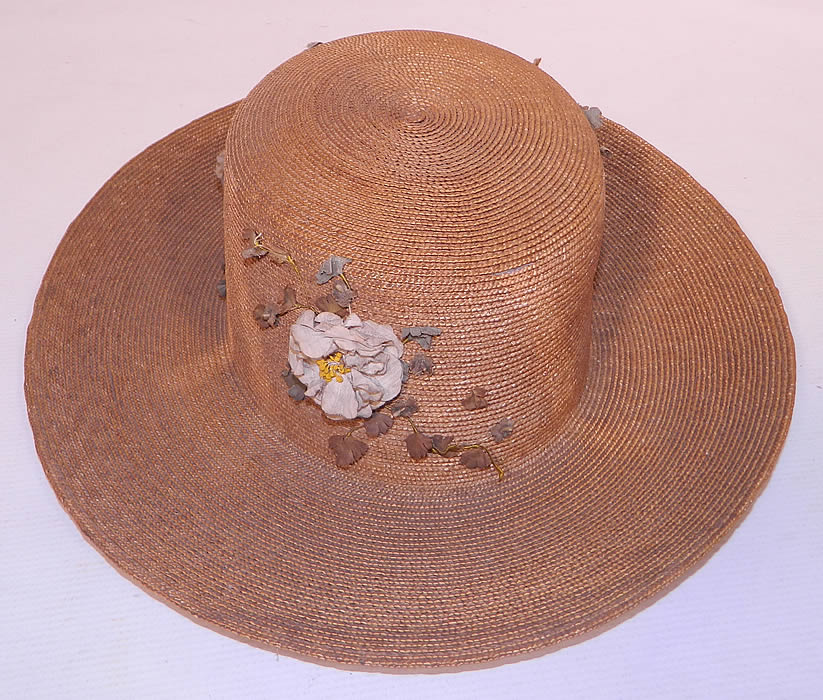 Edwardian Belle Epoque Natural Straw White Floral Wide Brim Boater Summer Hat. It is made of a tightly woven natural straw, with white silk flower trim and green leaf sprigs on the crown.
