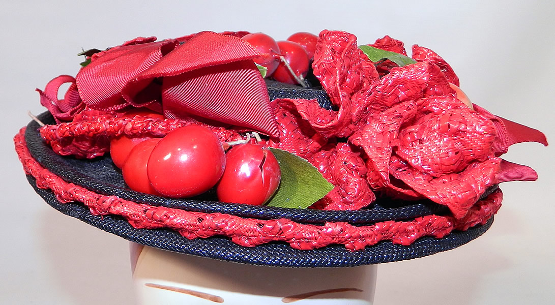 Vintage Red Straw Roses Cherries Small Skimmer Boater Tilt Hat Fascinator. It is made of a navy blue tight weave straw base, with red woven straw rose flowers, red plastic cherry fruit and a red silk grosgrain ribbon hatband bow trim.