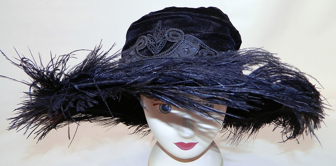 Edwardian Vintage Madame Gatecliff Modiste Black Velvet Lace Feather Trim Wide Brim Hat. It is made of a black velvet fabric, with black silk battenburg tape lace applique trim sewn around the crown and black ostrich feather plumes sewn around the brim.