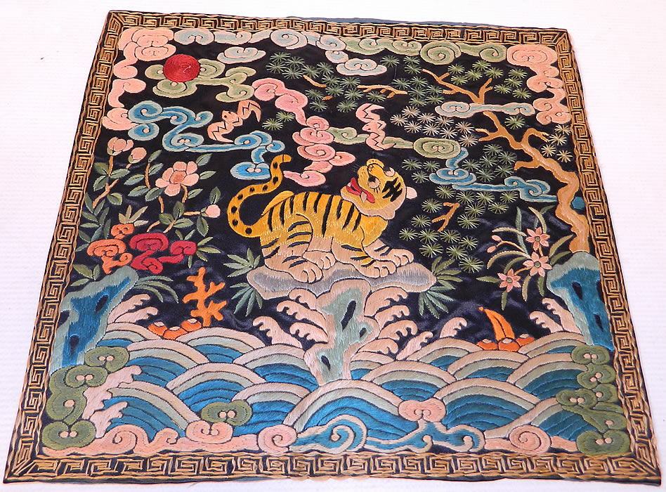 Antique Chinese Silk Embroidered Tiger Fourth Rank Military Badge Pair. There is a black stripe tiger with spotted long tail and open mouth in the center, with bats, trees, flowers and clouds of smoke in the background. There is a embroidered red sun disk on the left for a males badge and waves of water along the bottom.
